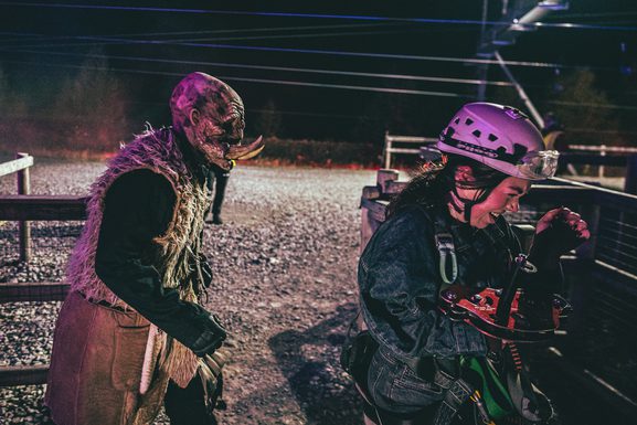 A tusked monster wearing a fur jacket chases after a screaming woman, who is wearing a zip line helmet, goggles and harness