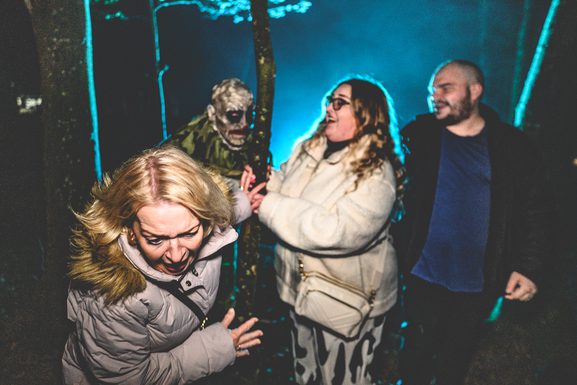 A clown lurks in in a woodland area lit with blue lighting, as a group of adults scream nearby