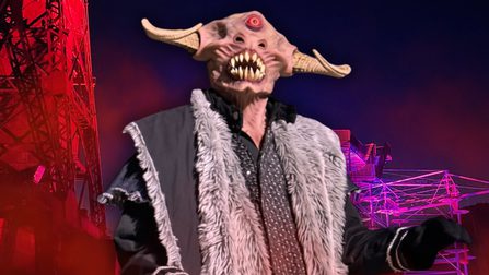 A horned creature with a red eye on its forehead, wearing a fur coat, looks down on the camera whilst standing in front of red-illuminated adventure structures.
