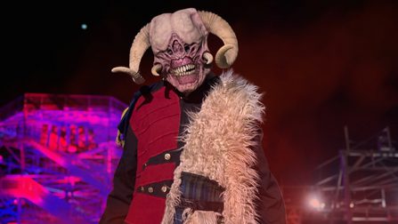 A horned monster with small slitted eyes wearing a fur coat stands in front of a large, brightly-illuminated climbing course.