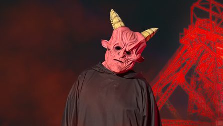 A red-faced and horned monster stares at the camera, surrounded by red mist. A tower mining structure illuminated in red stands behind the monster.