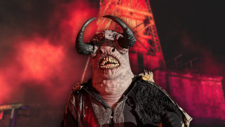 A horned monster with sharp teeth and small slitted eyes stands in front of a red mining tower structure, illuminated in eerie red lighting